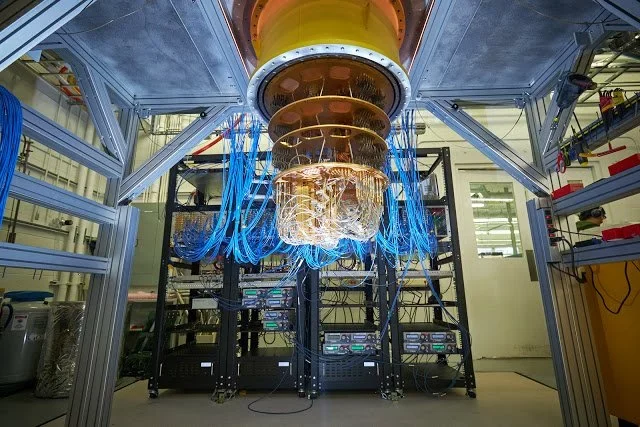 A quantum computing rack in a laboratory, its gold-plated cooling stages hung with dense bundles of blue cabling.