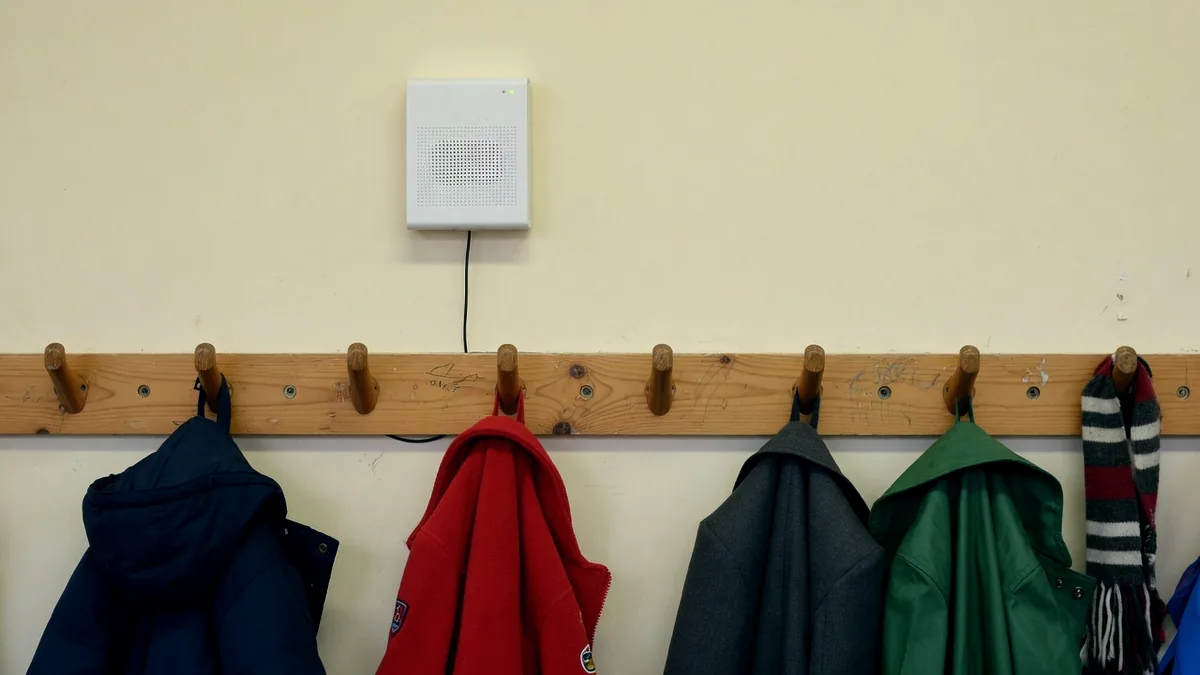 A small air quality sensor on a classroom wall above a row of coat pegs hung with coats
